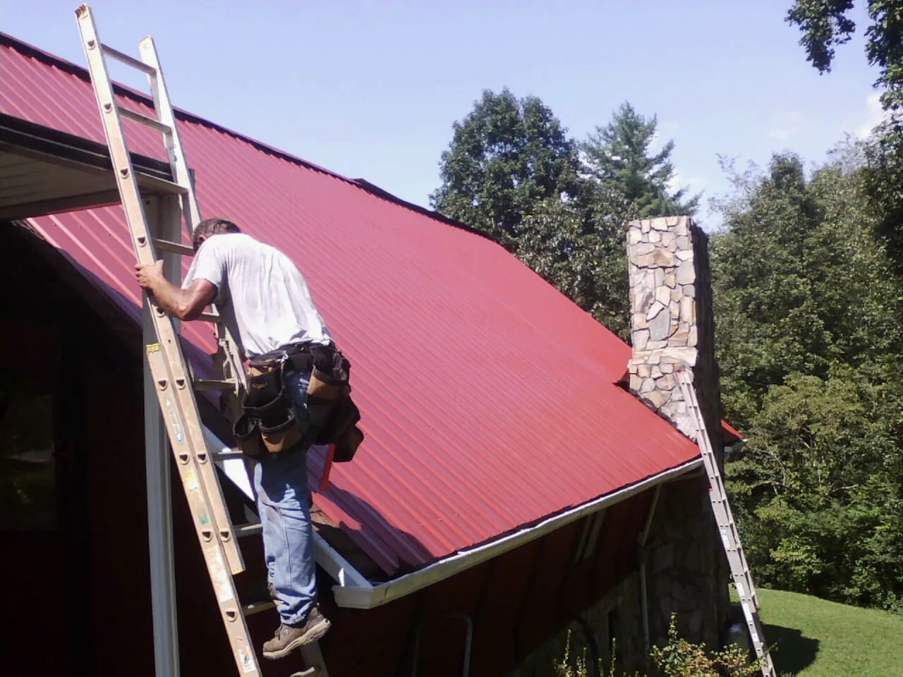 Roofer on a ladder beside a red metal roof and stone chimney — Soffit & Fascia Repair by Roofing Brothers in Baker City